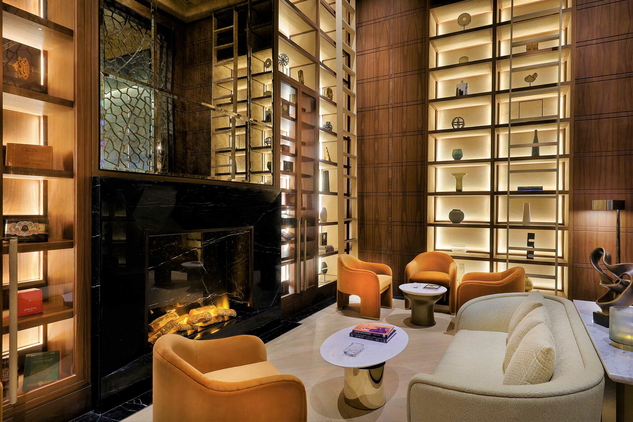 Cozy modern hotel lounge with amber chairs, a beige sofa, lit bookshelves, and a black stone fireplace. Soft lighting creates a warm, inviting atmosphere.