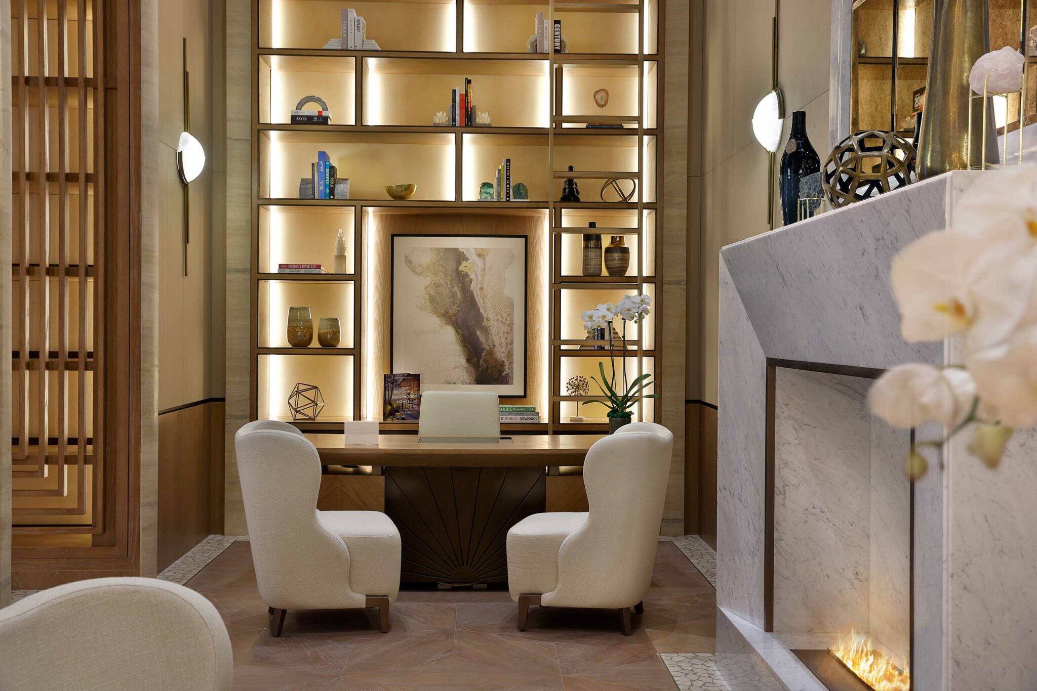 Elegant hotel study area with a warm ambiance. Features a bookshelf with art, decor items, and backlighting. Two cozy chairs face a wooden desk, near a lit fireplace.