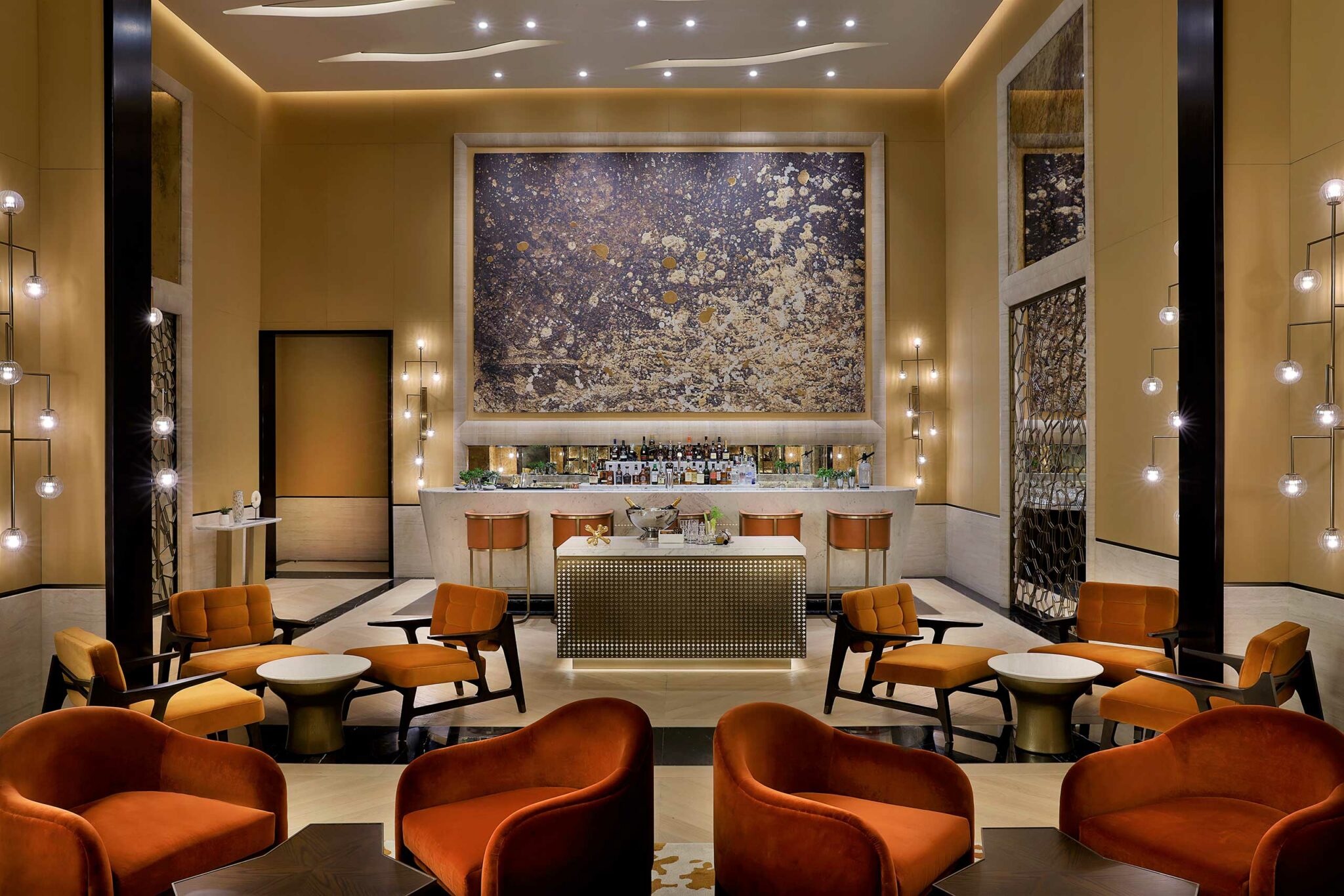 Elegant hotel lounge with warm lighting, modern orange chairs, round tables, and a well-stocked bar. A large abstract painting adorns the wall. The atmosphere is sophisticated and inviting.