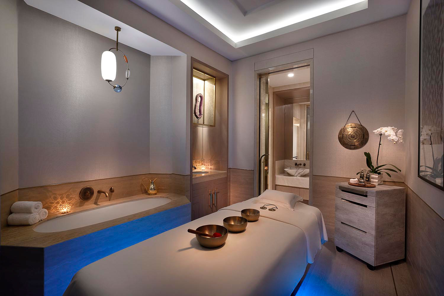 A serene hotel spa treatment room with a massage bed, warm lighting, and a filled bathtub. Bowls on the table and an orchid create a calming atmosphere.