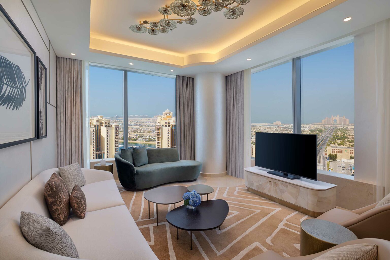 Elegant modern suite living room with large windows showing a cityscape. Features a sleek TV, plush seating, abstract art, and a stylish chandelier.