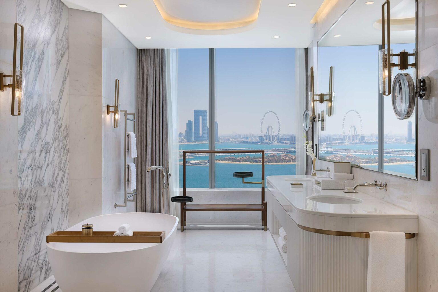 Luxurious suite bathroom with a large marble bathtub, elegant fixtures, and a panoramic window view of a city skyline and ocean, creating a serene atmosphere.