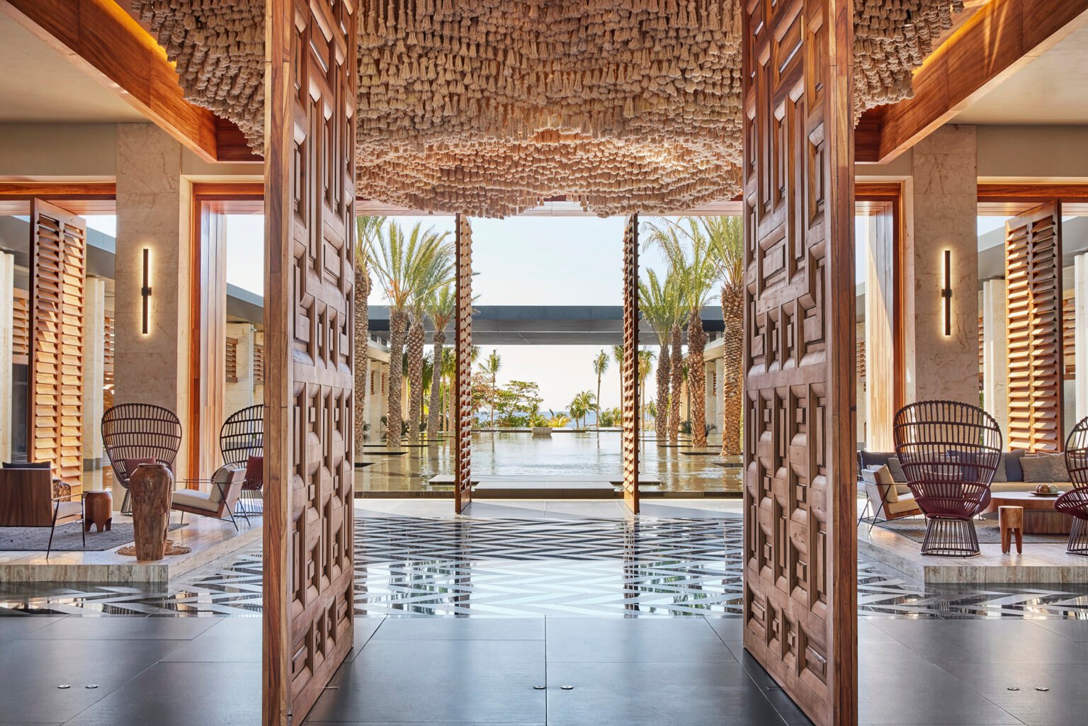 Luxurious open-air hotel lobby with intricate wooden doors, modern seating, and palm trees surrounding a reflective pool, creating a serene, inviting atmosphere.
