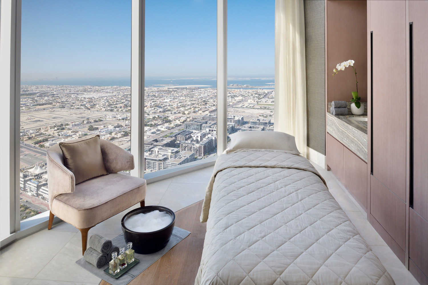 A serene spa treatment room with a massage bed and a plush chair, featuring large windows that offer panoramic views of Dubai, creating a tranquil and luxurious relaxation space.