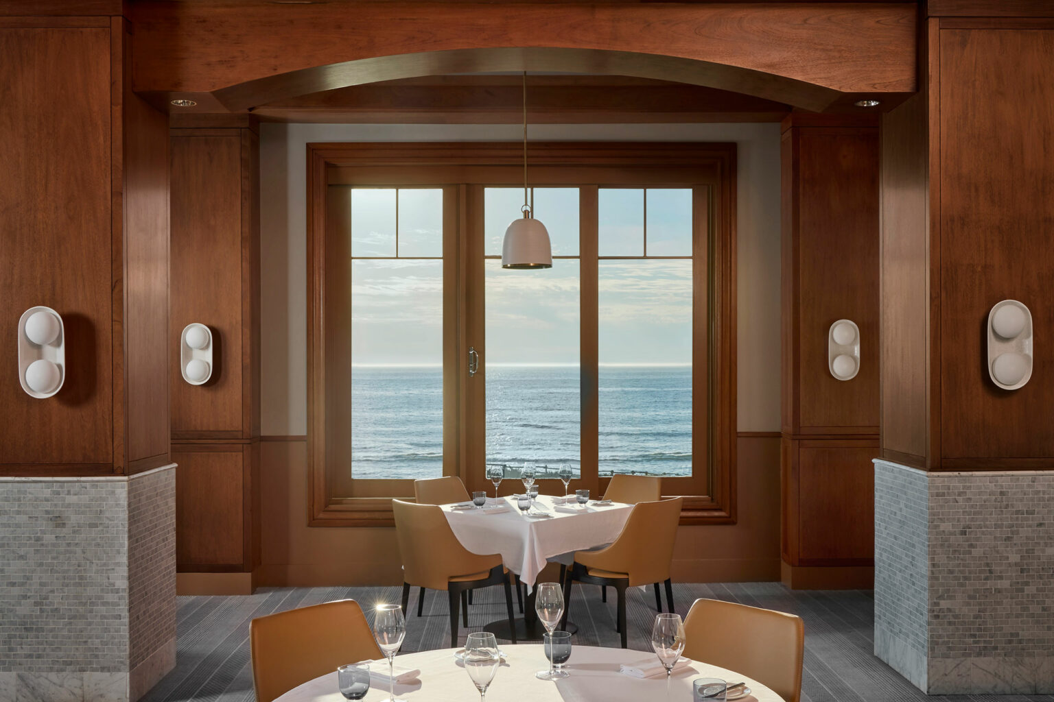 Hotel restaurant interior with wooden decor featuring a table set for four with tan chairs. A large window offers a serene ocean view under a softly lit pendant lamp.
