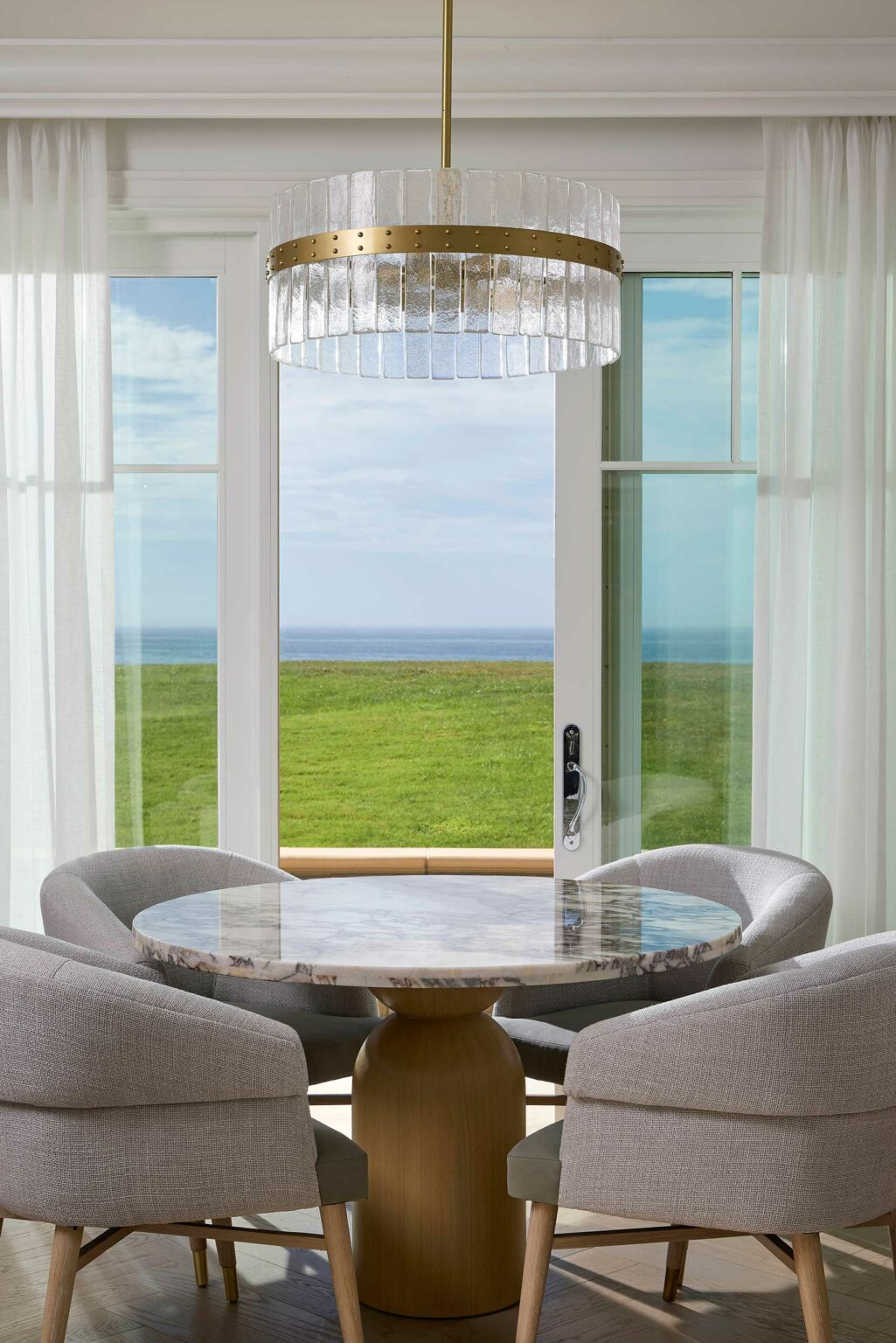 A cozy, elegant hotel dining area features a round marble table, two plush gray chairs, and a large glass chandelier. Sunlit French doors reveal a lush green lawn and tranquil ocean view, evoking a serene, inviting atmosphere.