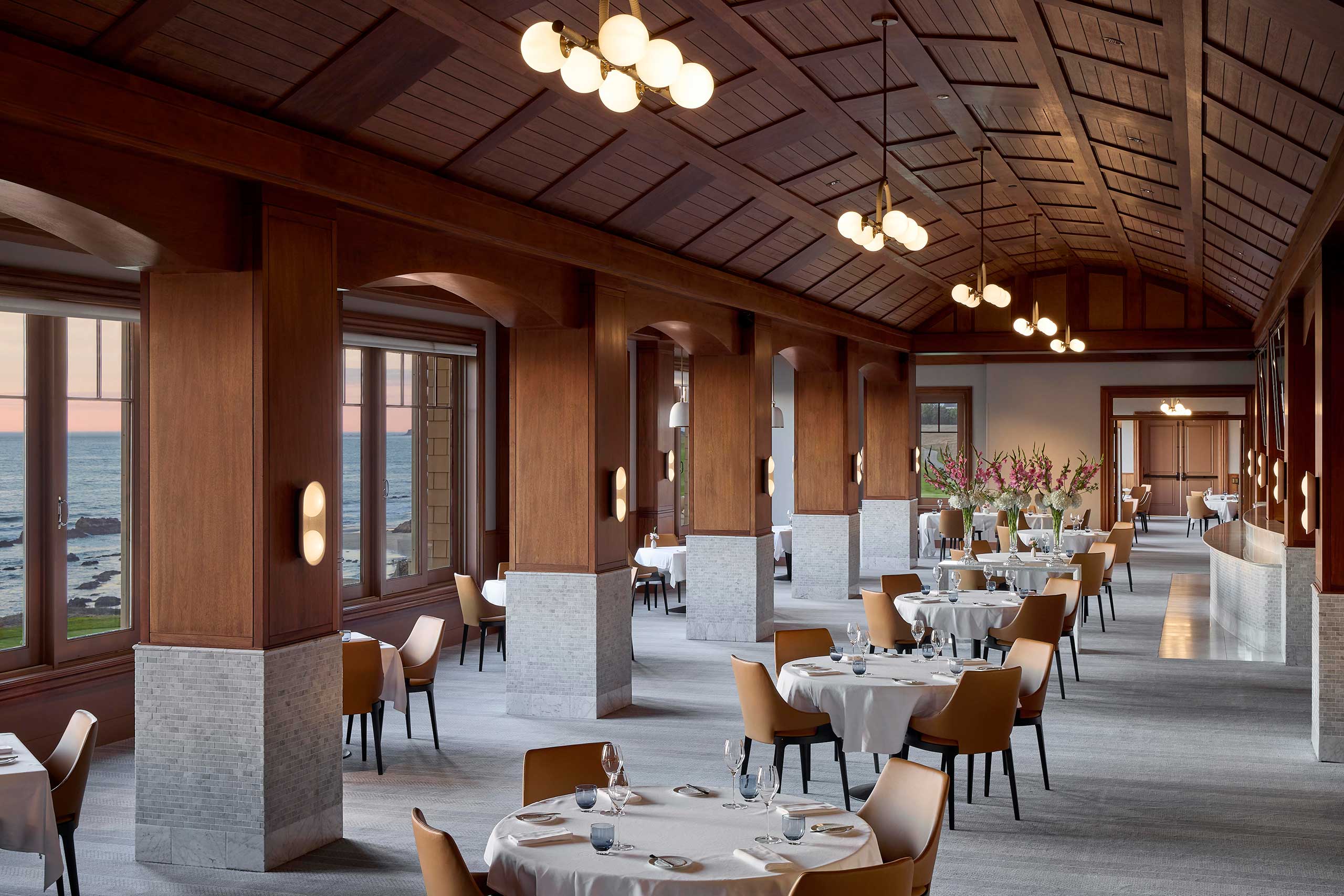 Elegant hotel restaurant with wooden beams, round tables, and leather chairs. Large windows offer an ocean view. Soft lighting gives a warm, inviting ambiance.