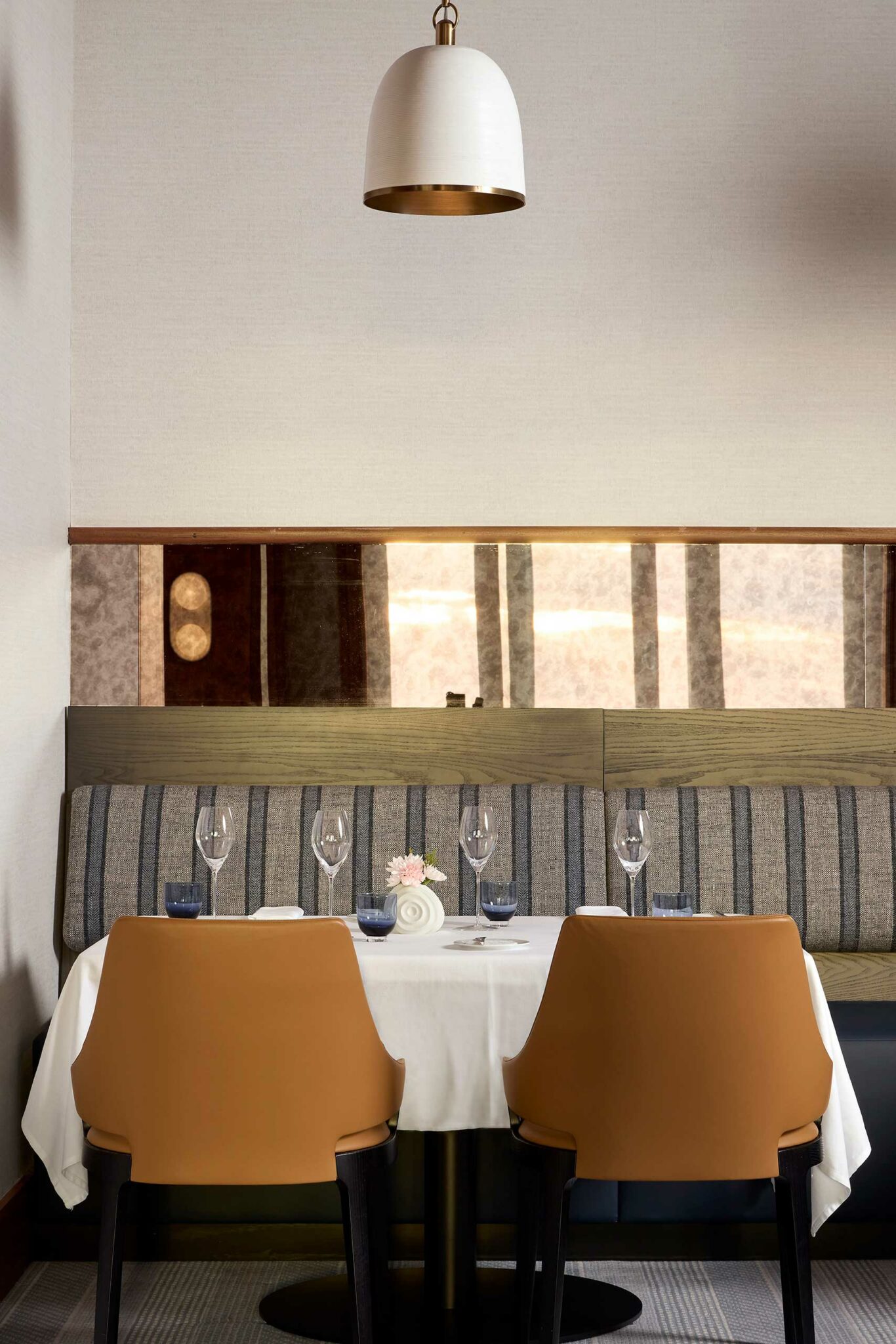 Modern hotel restaurant setting with a table set for four, featuring a white tablecloth, elegant wine glasses, and a small flower centerpiece. Brown leather chairs face striped banquette seating under a chic white pendant lamp.