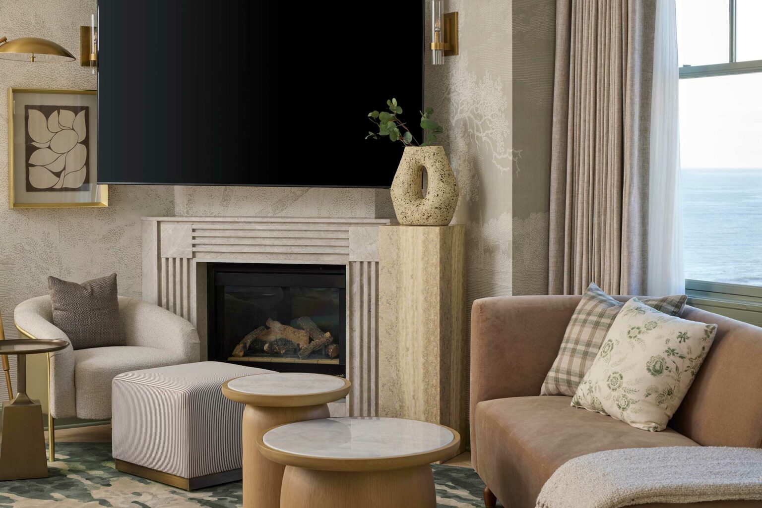 Elegant hotel lounge with a modern fireplace, large TV, and comfortable seating. Neutral tones, patterned pillows, and an ocean view create a serene ambiance.