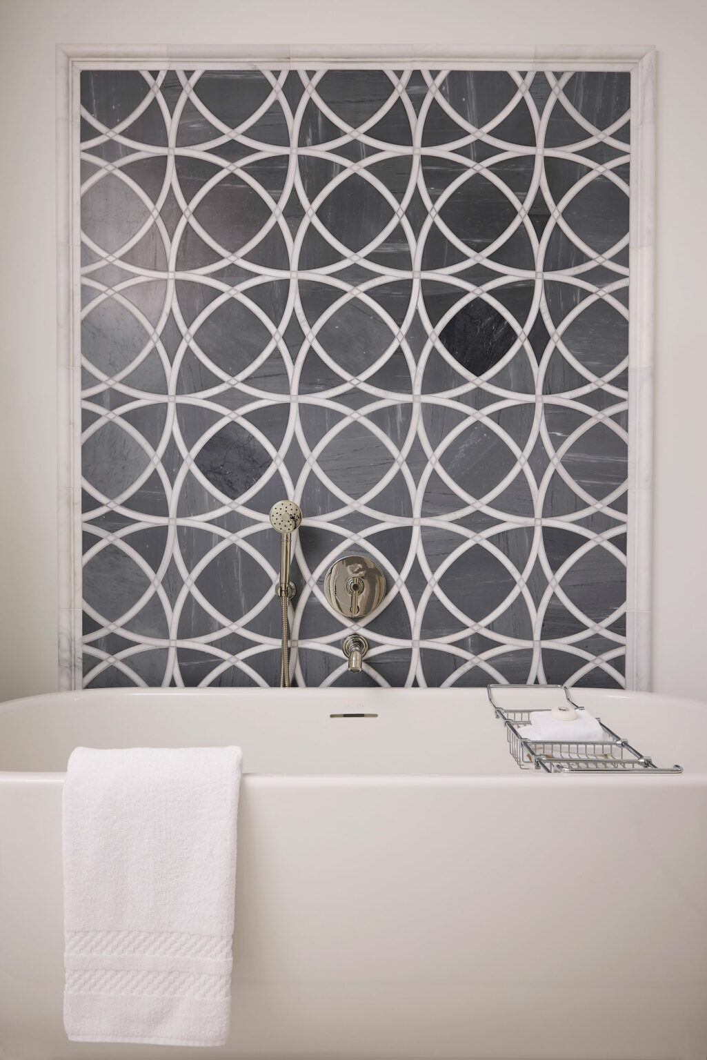 Modern bathroom with a white bathtub in front of a dark geometric tile wall with overlapping circles. A towel hangs over the tub, creating a serene ambiance.