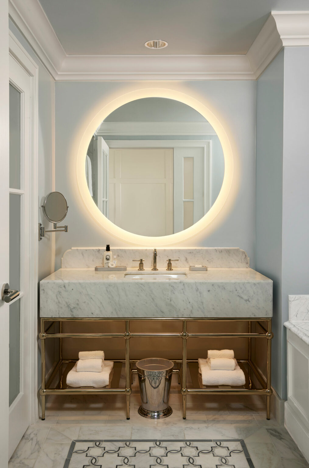 Elegant suite bathroom with a round, backlit mirror above a marble vanity. Brass fixtures, neatly folded towels, and a metallic bin evoke a modern, serene ambiance.