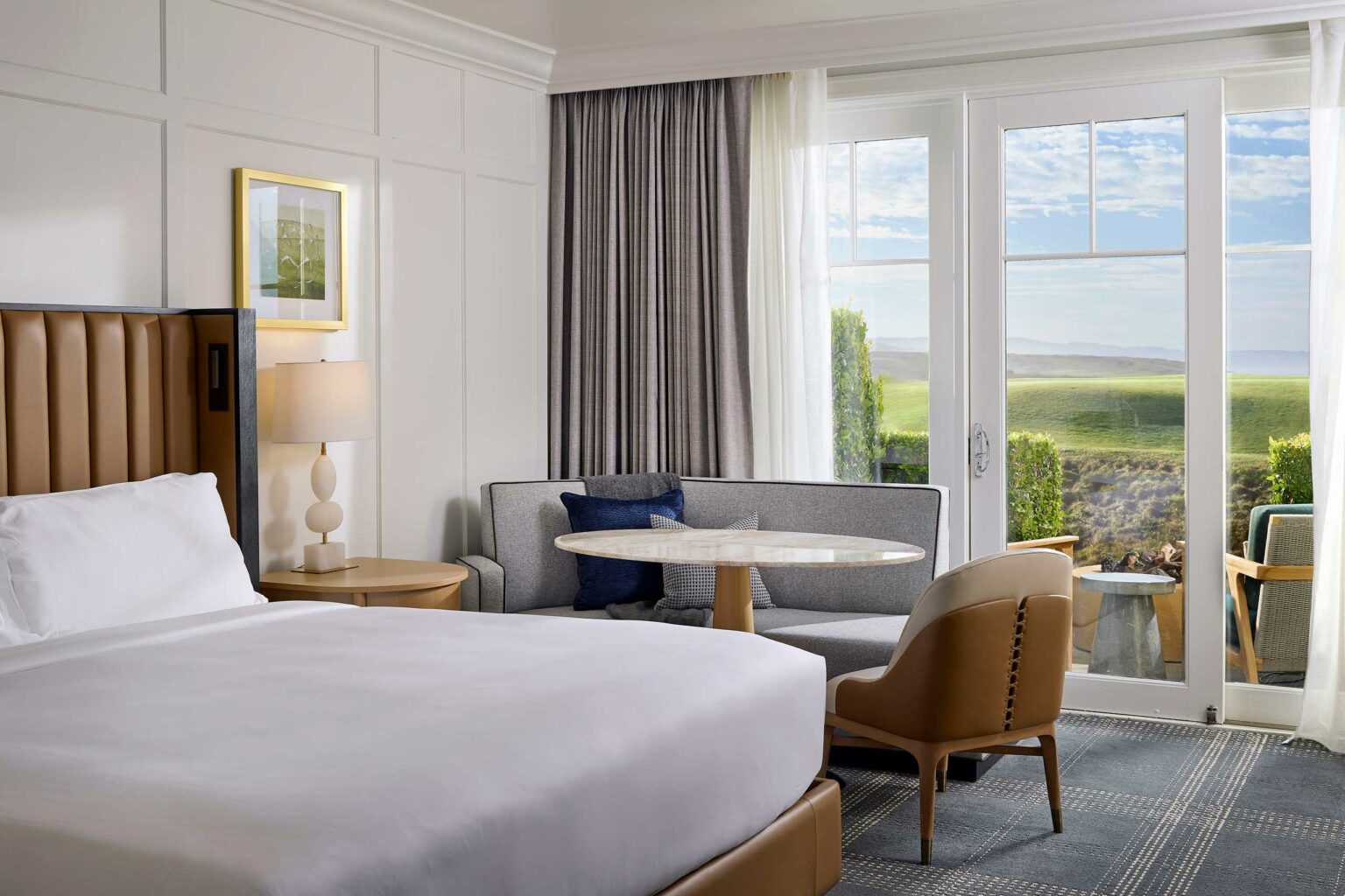 Elegant hotel room with a plush bed, modern lamp, and cozy seating area. Large windows offer a tranquil view of greenery under a blue sky.