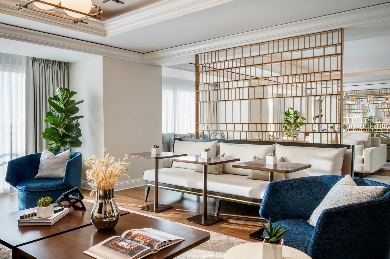 Bright, modern hotel lounge with white sofa, blue armchairs, and wood tables. Large plant, decorative vases, and grid-like partition add elegance.