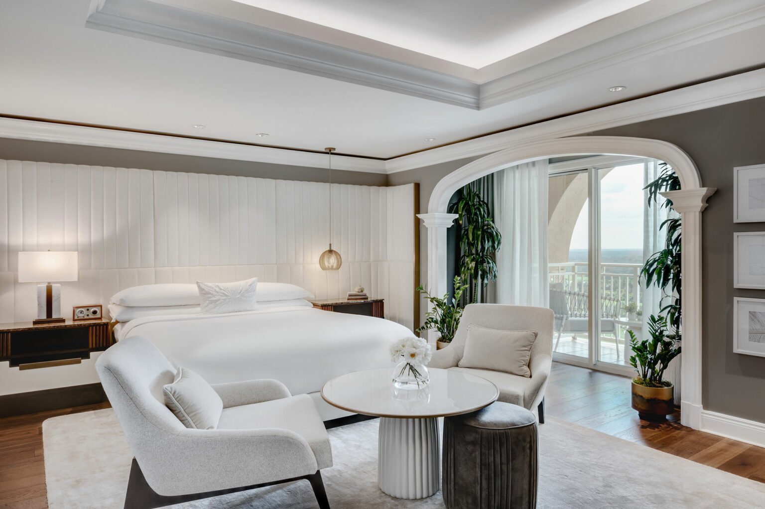 Luxurious suite bedroom with a king-sized bed, elegant white decor, cushioned chairs around a small table, and large windows opening to a balcony view.