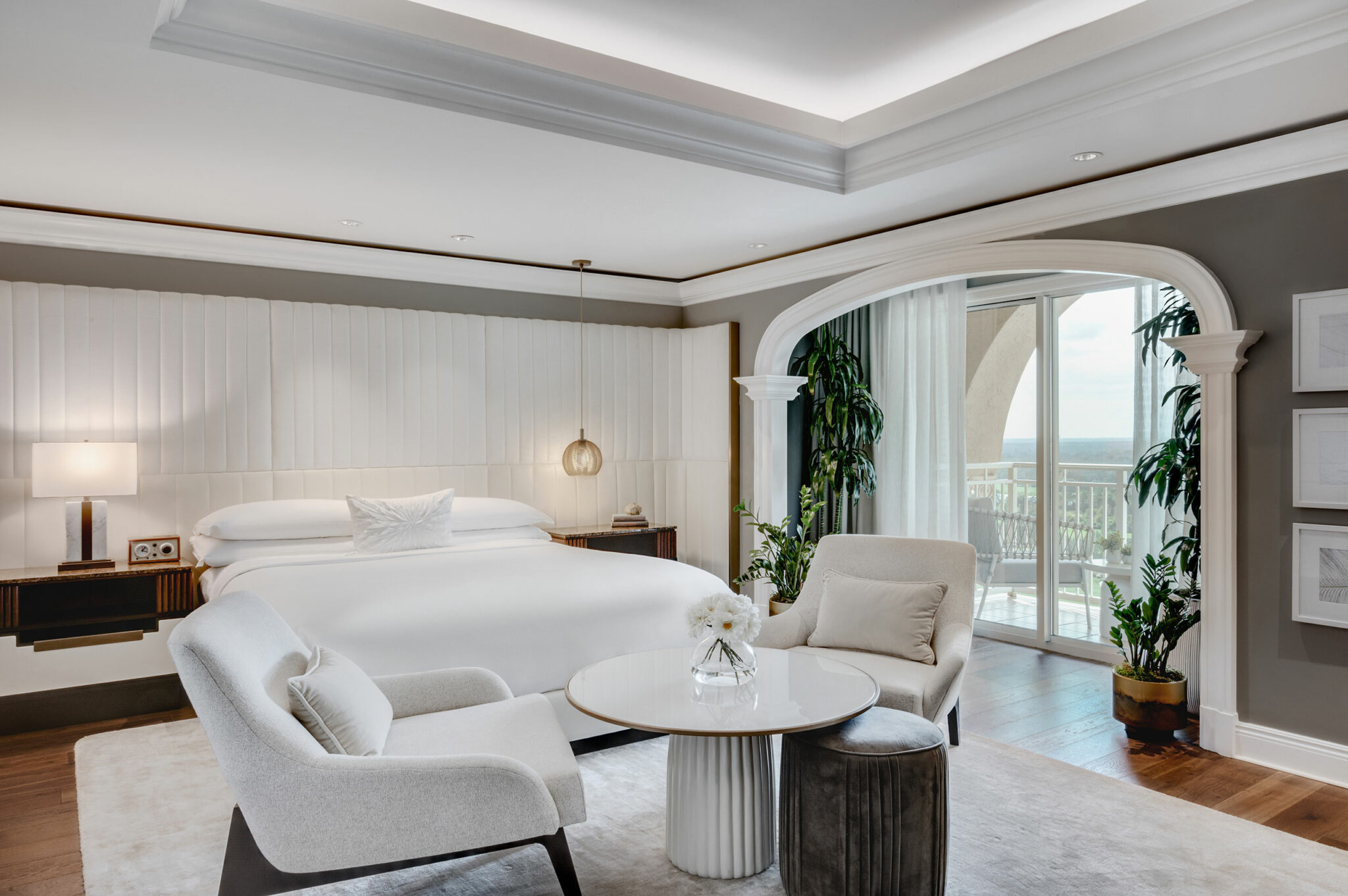 Luxurious suite bedroom with a king-sized bed, elegant white decor, cushioned chairs around a small table, and large windows opening to a balcony view.