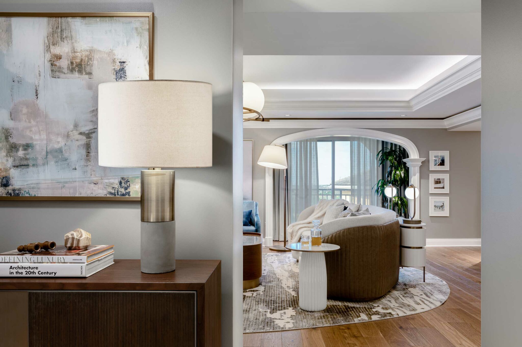 Modern suite living room with muted tones, featuring a wooden side table with a lamp, abstract art, curved sofa, large window, and green plants. Cozy and elegant.