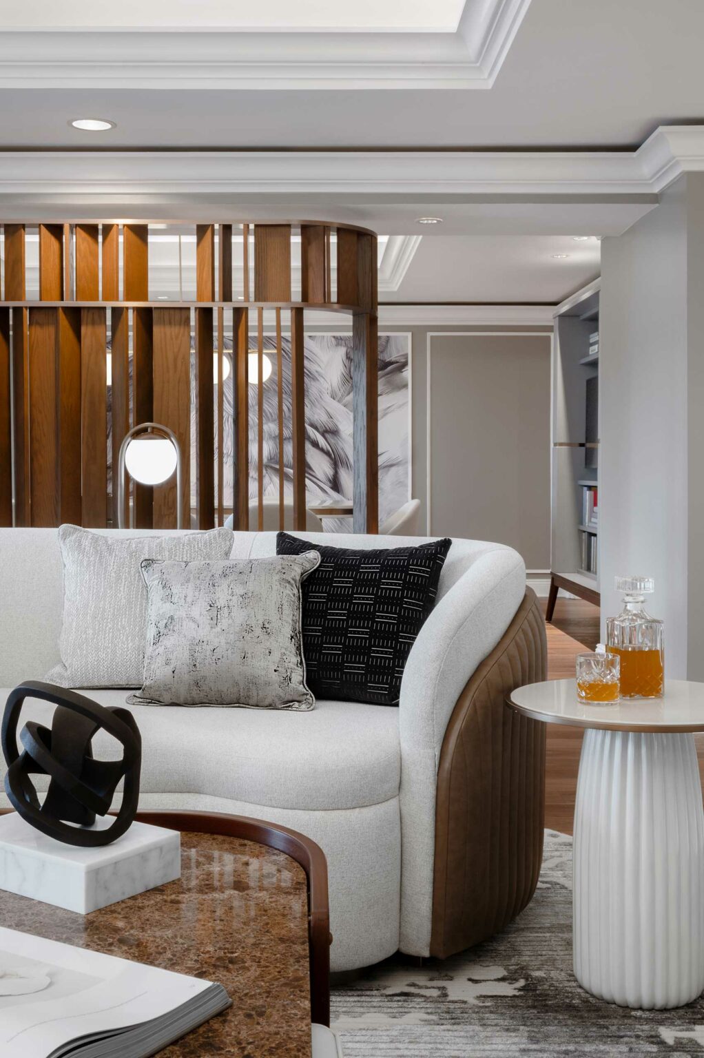 A modern suite living room features a curved white sofa with patterned cushions, a wooden accent wall, stylish decor, and a table with crystal glasses. The atmosphere is elegant and cozy.