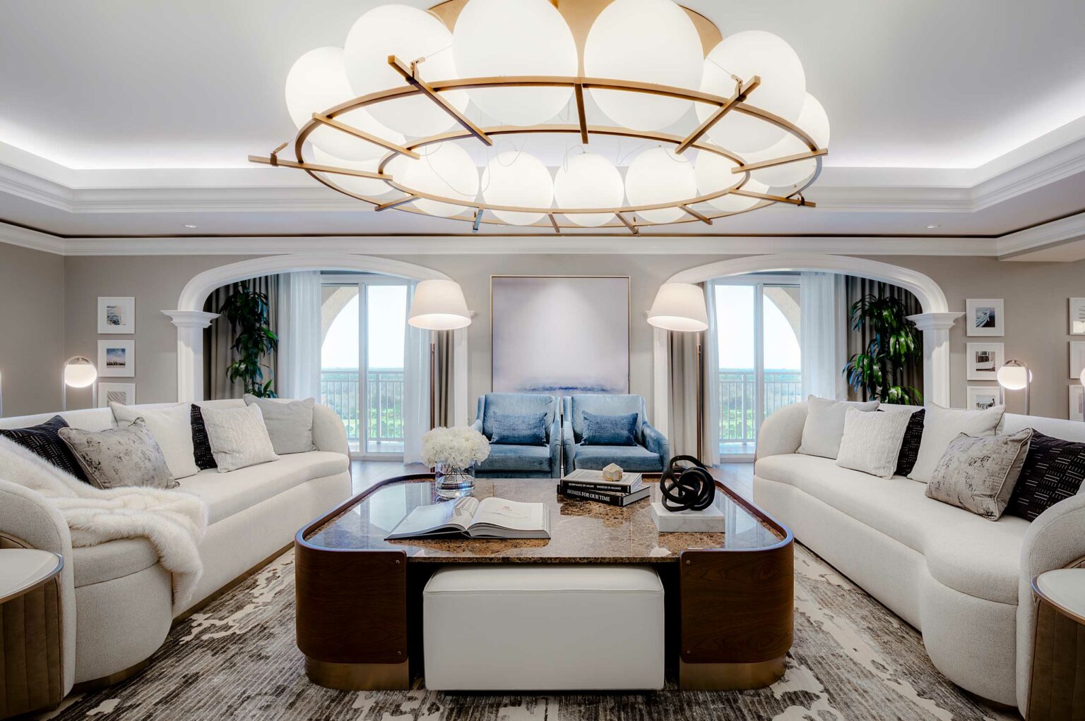 Sophisticated suite living room with white sofas, blue armchairs, a large chandelier, and a marble coffee table. Two glass doors lead to a balcony, creating an elegant and serene ambiance.