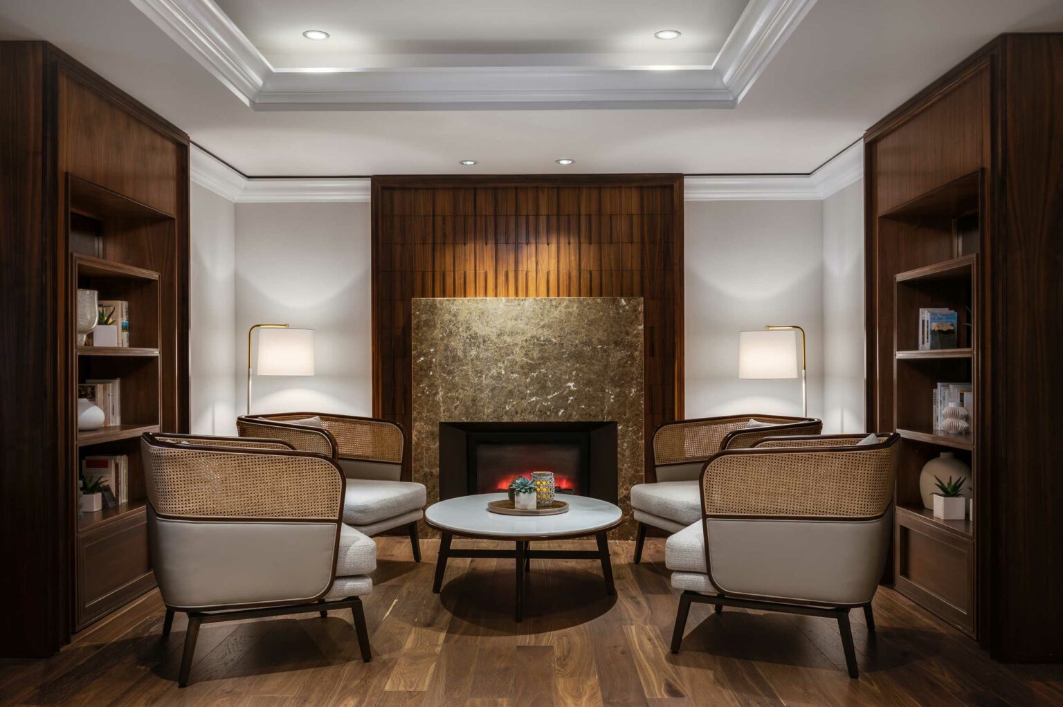 Cozy modern hotel lounge with four elegant chairs around a circular table, set in front of a fireplace. Warm lighting and bookshelves create an inviting ambiance.