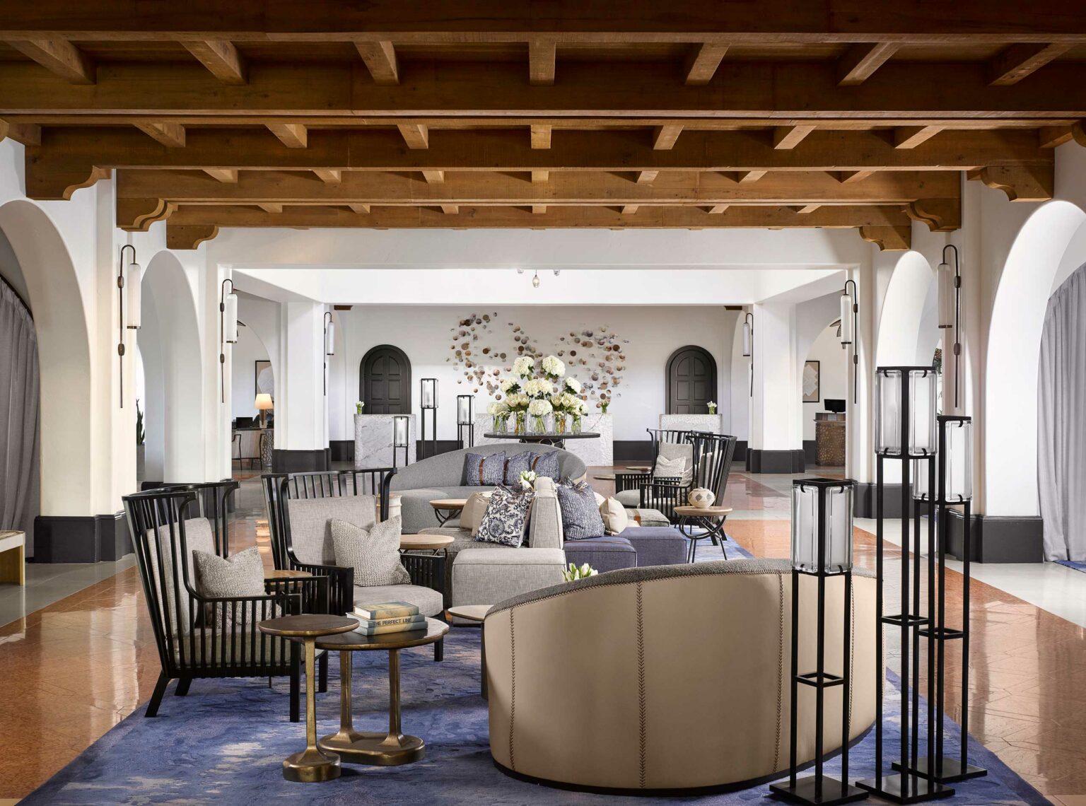 Spacious hotel lobby with elegant, modern furnishings including plush sofas, armchairs, and a floral centerpiece. Warm tones and soft lighting create a welcoming atmosphere.