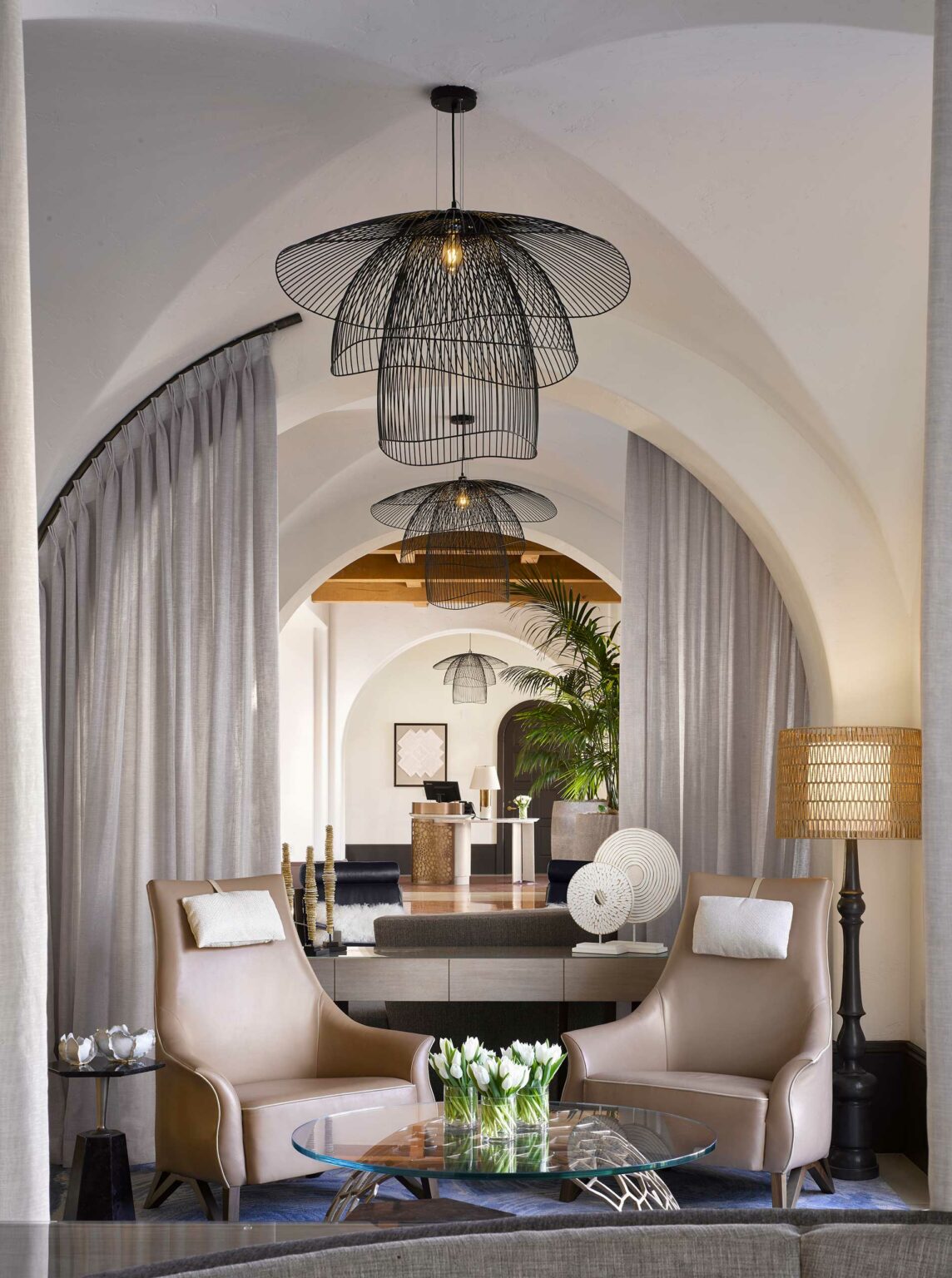 Elegant modern hotel lounge with arched ceilings and artistic pendant lights. Two tan chairs face a glass table with white tulips. Neutral tones.