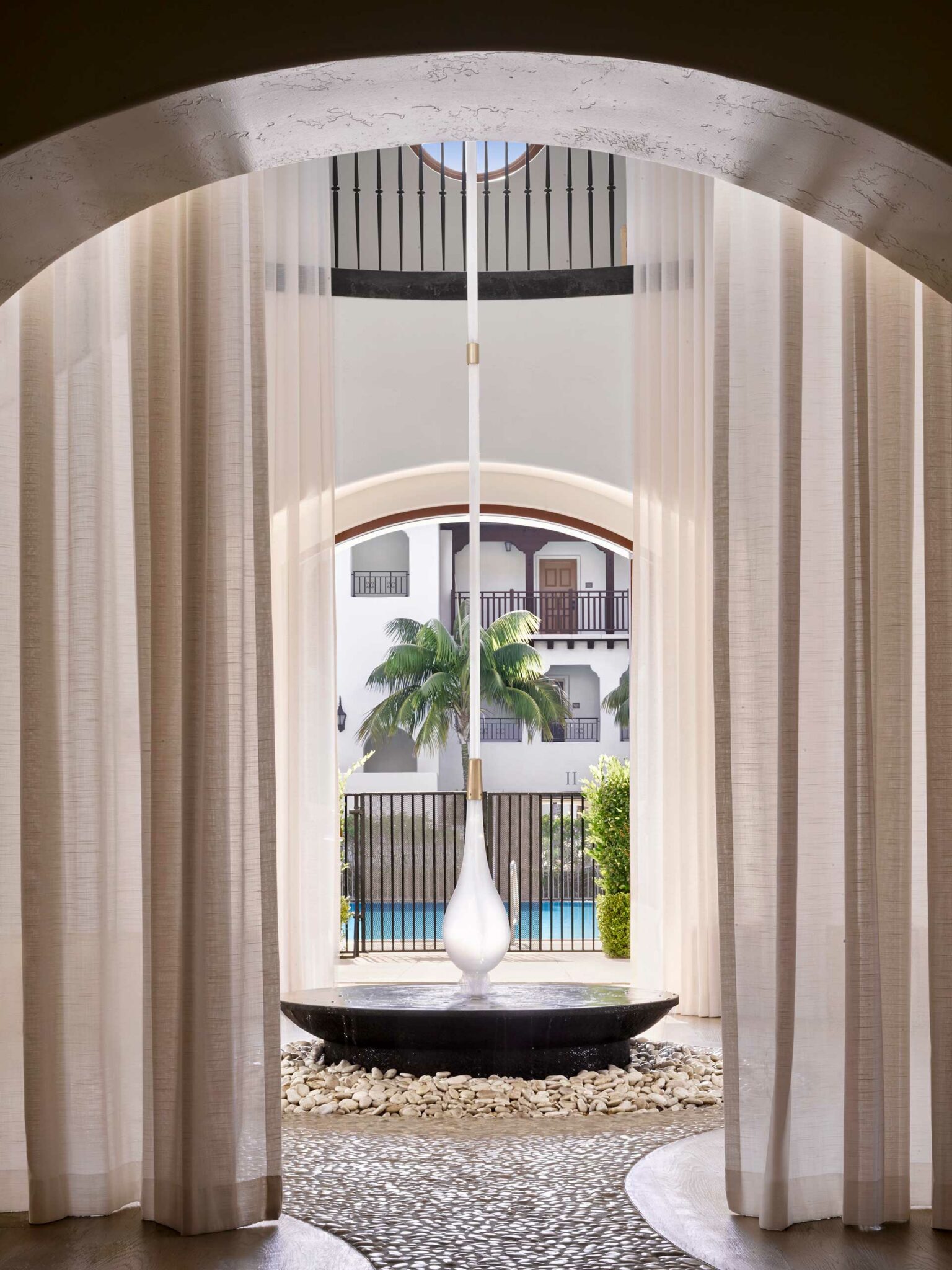 Elegant hotel lobby with tall curtains framing a modern, teardrop-shaped light above a circular fountain. Beyond the window, palm trees and a pool.