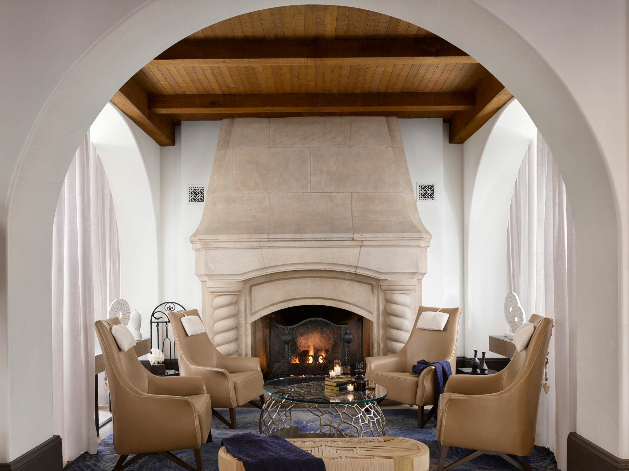Elegant hotel lounge nook with a large stone fireplace, four tan armchairs, and a glass coffee table. A cozy fire burns, creating a warm, inviting atmosphere.