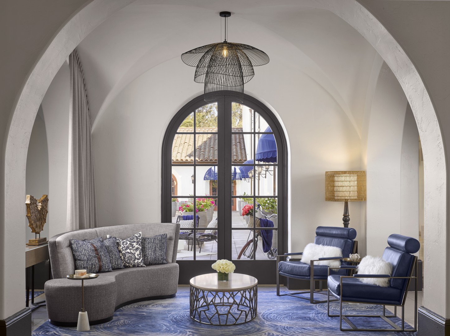 Sophisticated hotel lounge nook with arched window, blue chairs, gray sofa, and flower centerpiece. Natural light, elegant ambiance, modern decor.