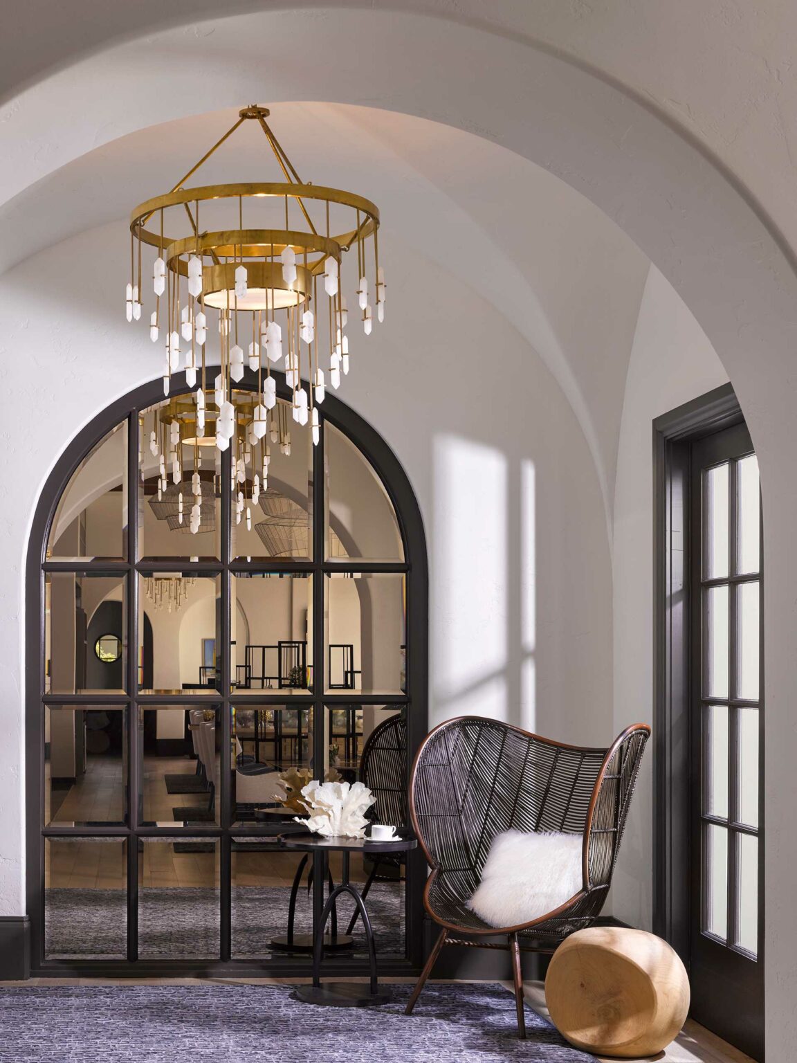 Luxury hotel lounge corner featuring an arched window with reflection of a chandelier, seating area with wicker chair and side table. Soft light creates a serene, modern ambiance.