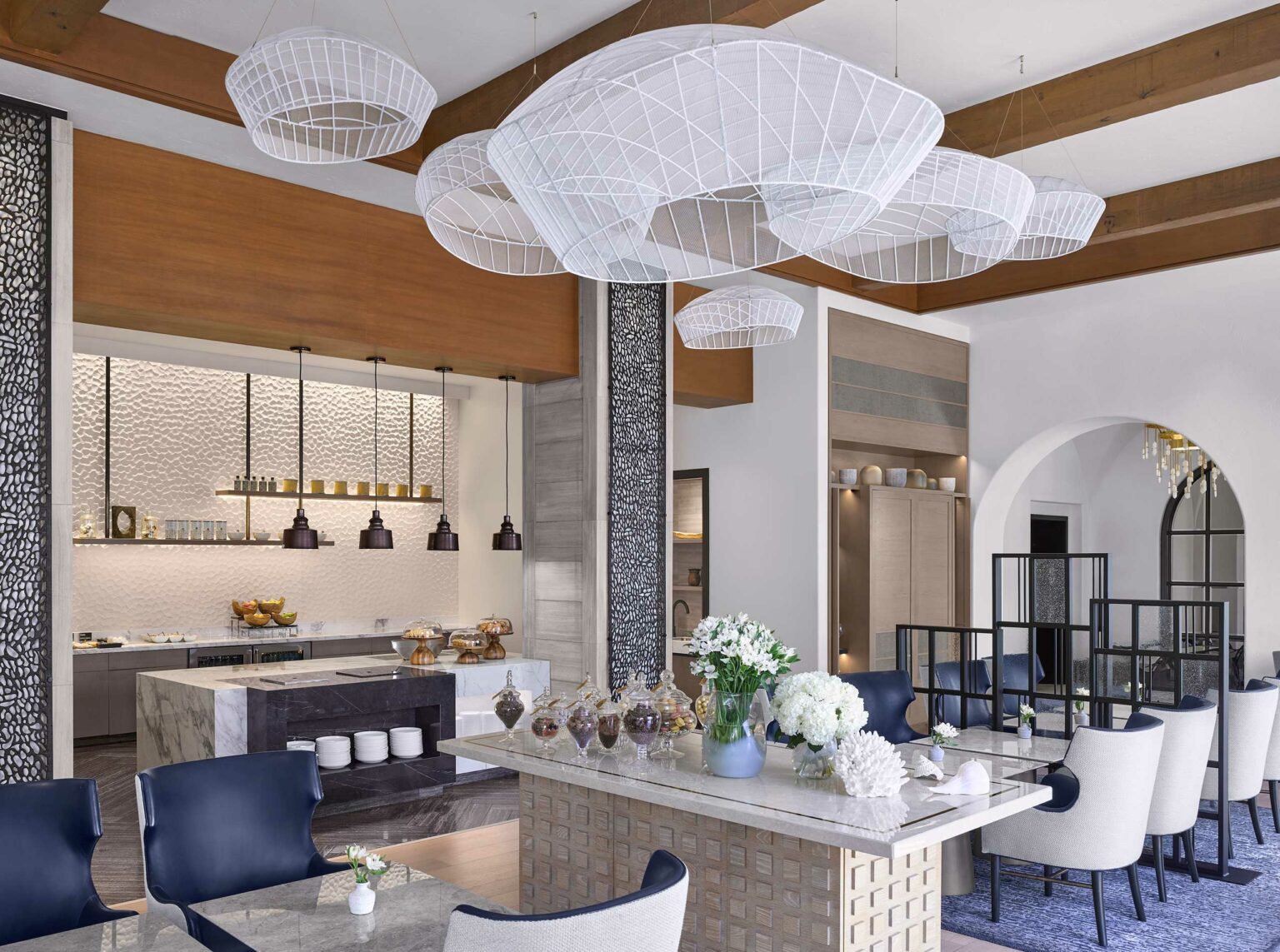 Modern hotel dining area with a large, white abstract chandelier hanging from a wood-beamed ceiling. Contemporary decor includes blue chairs, a marble table set with white flowers. There are open shelves, arches, and stylish lighting, creating an elegant and inviting atmosphere.