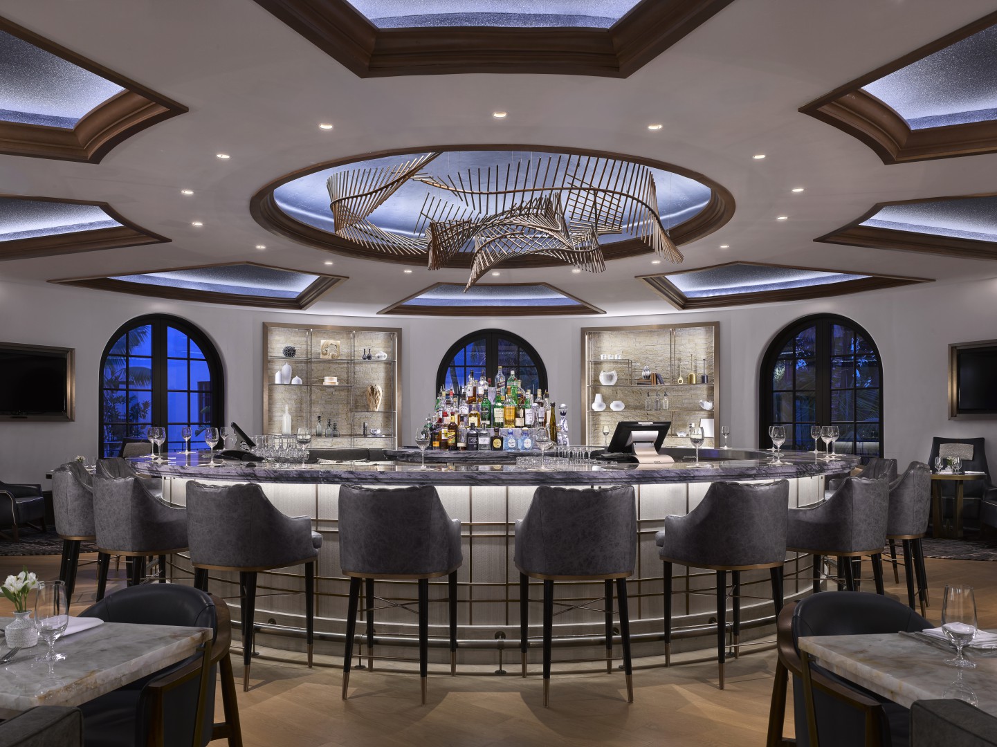 A stylish, modern hotel bar with a sleek circular counter, elegant gray stools, and a central liquor display. Artistic ceiling lights create a luxurious ambiance.