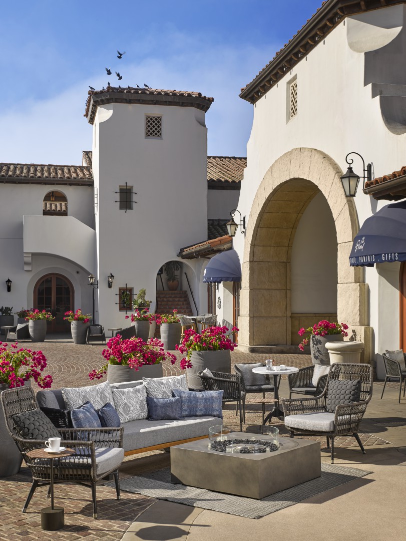 Outdoor courtyard of a luxury hotel with Spanish-style architecture featuring white walls and archways. Cozy seating with cushions and vibrant pink flowers add warmth.