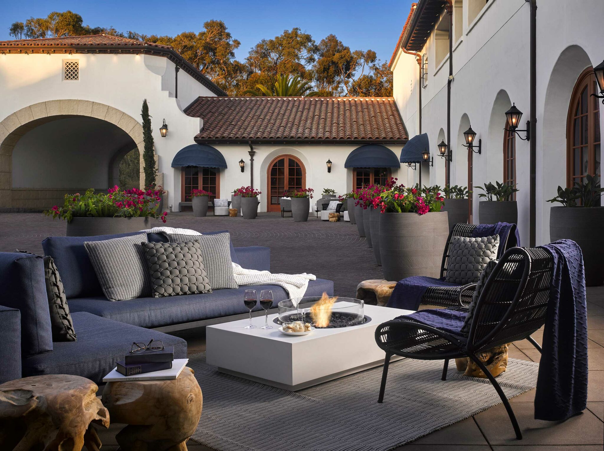Outdoor patio of a luxury hotel with cozy grey and blue seating around a white fire pit table. Wine glasses and vibrant pink flowers evoke a relaxing, elegant atmosphere.