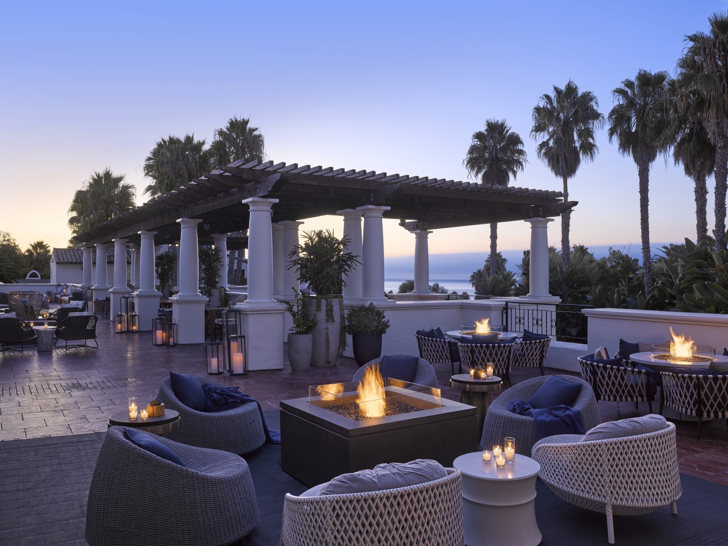 A serene outdoor patio of a luxury hotel at sunset with cozy seating around fire pits, surrounded by palm trees and overlooking the ocean. The ambiance is tranquil and inviting.