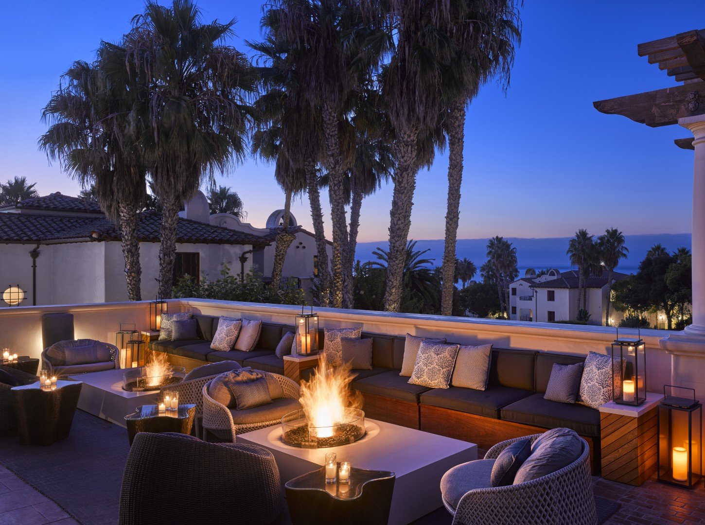 Luxurious outdoor patio of a luxury hotel at twilight, featuring cozy seating around a warm fire pit. Palm trees and a calm ocean backdrop add to the serene ambiance.