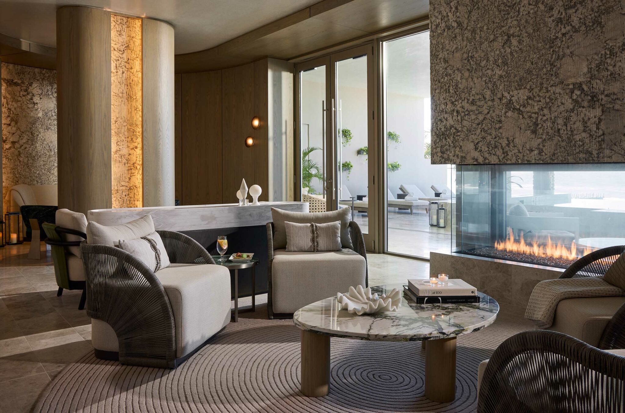 Modern hotel lounge with beige and gray armchairs around a marble table, a glass fireplace, and large windows. Relaxed ambiance with soft lighting.