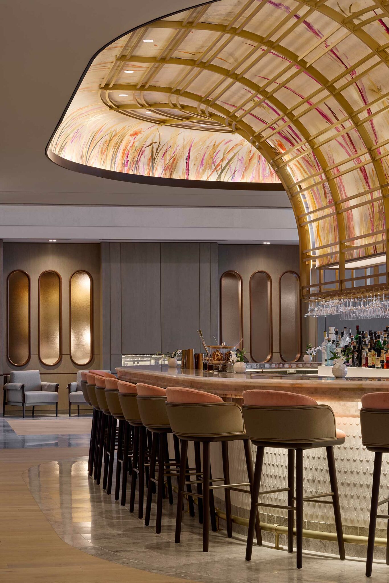 Elegant bar corner of a hotel lounge with a curved, golden canopy and floral patterns. Plush pink chairs line the sleek bar, exuding a luxurious and inviting atmosphere.