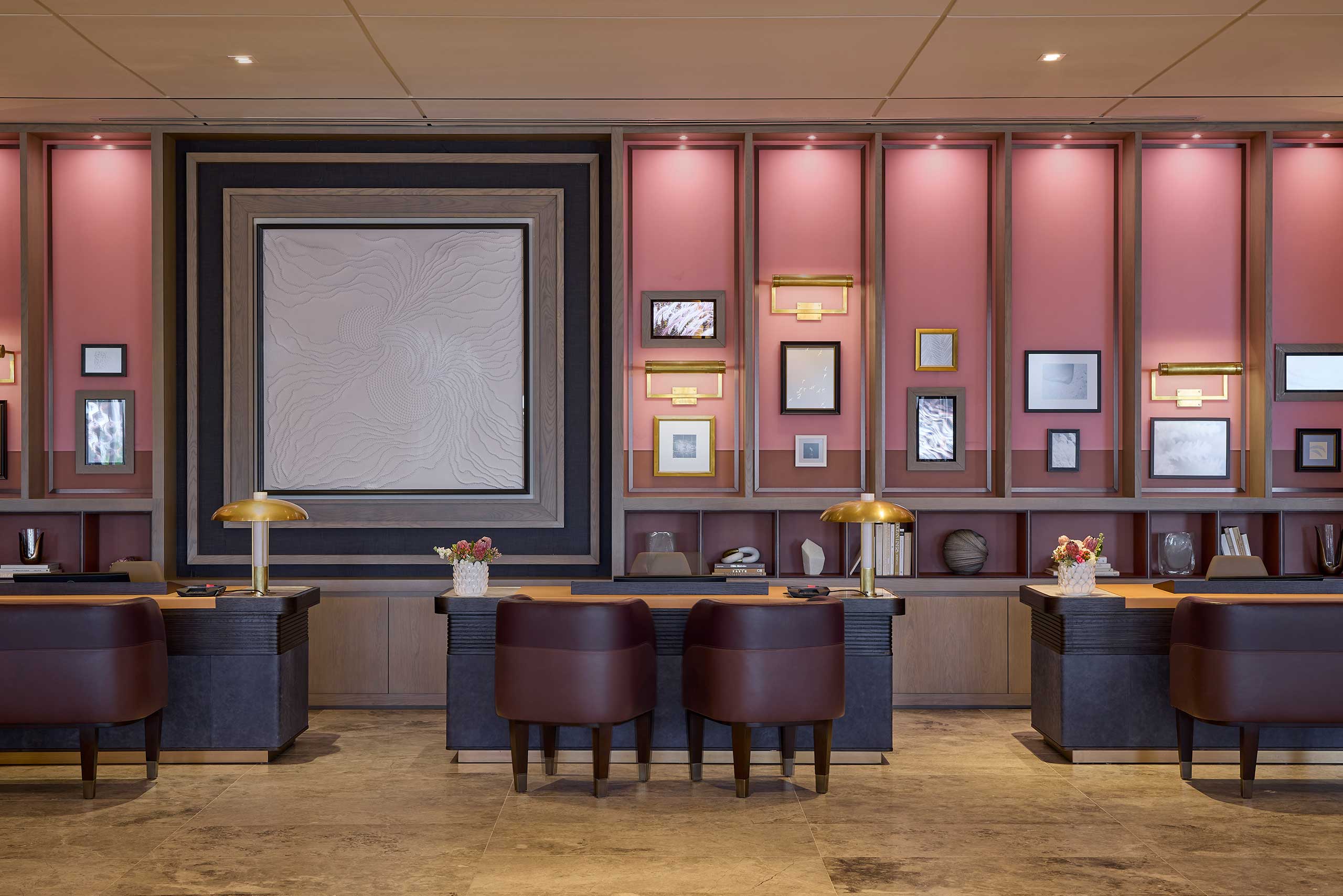 A stylish hotel lobby with pink walls adorned with framed art. Dark wood desks feature brass lamps and brown chairs, exuding a warm, inviting atmosphere.