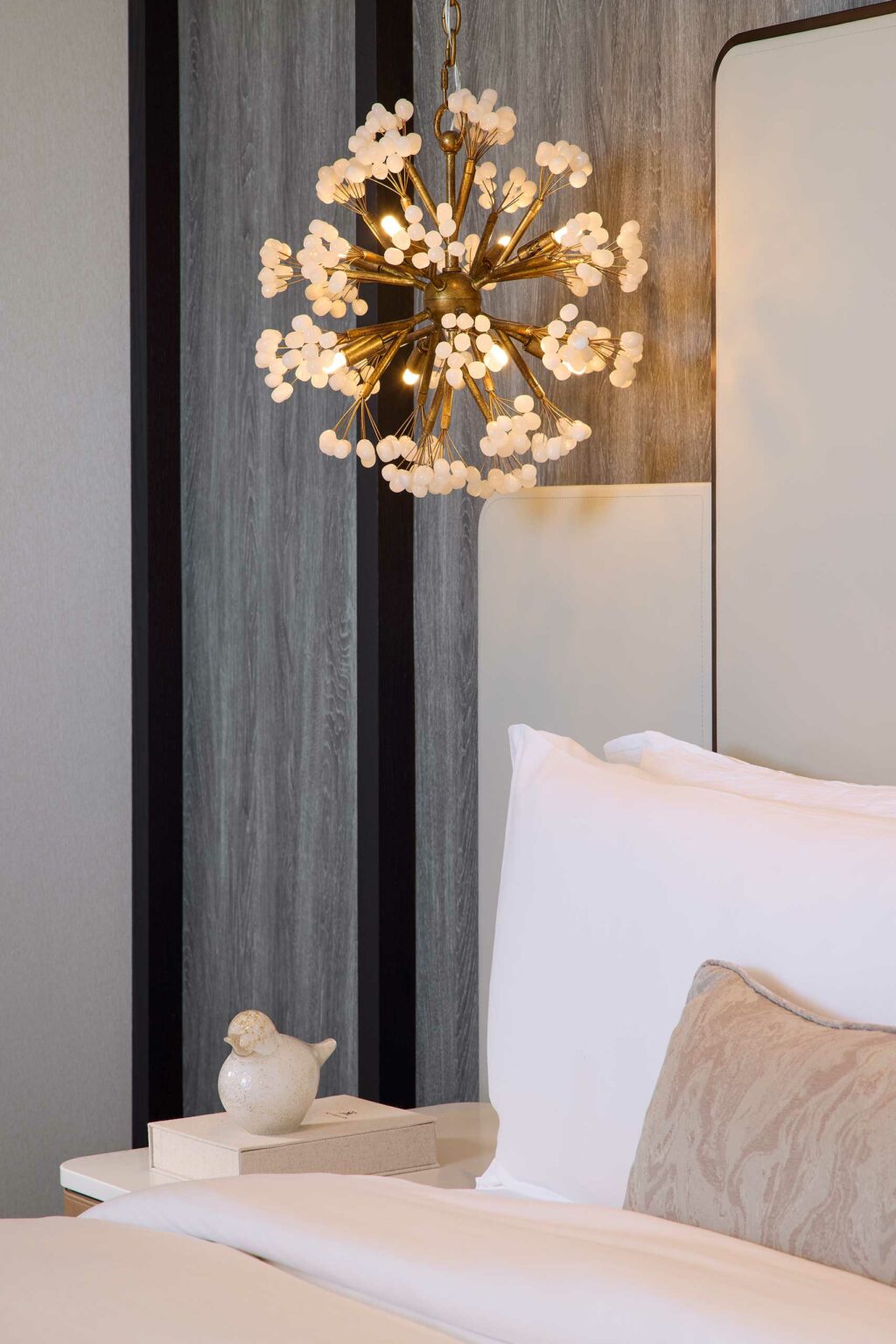 Elegant suite bedroom scene with a bright, modern chandelier shaped like a starburst, white and beige bed linens, and a decorative bird figurine on the nightstand.