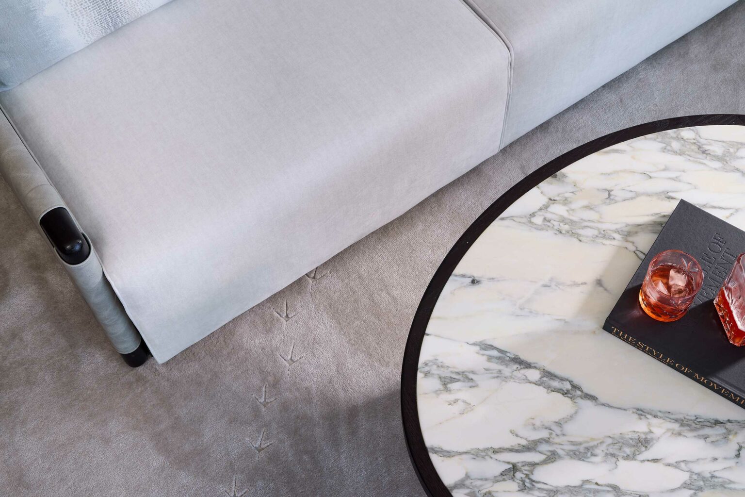 A gray couch meets a round marble coffee table on a carpeted floor. The table holds a book and a reddish drink, creating a calm, modern atmosphere within a luxury hotel suite setting.