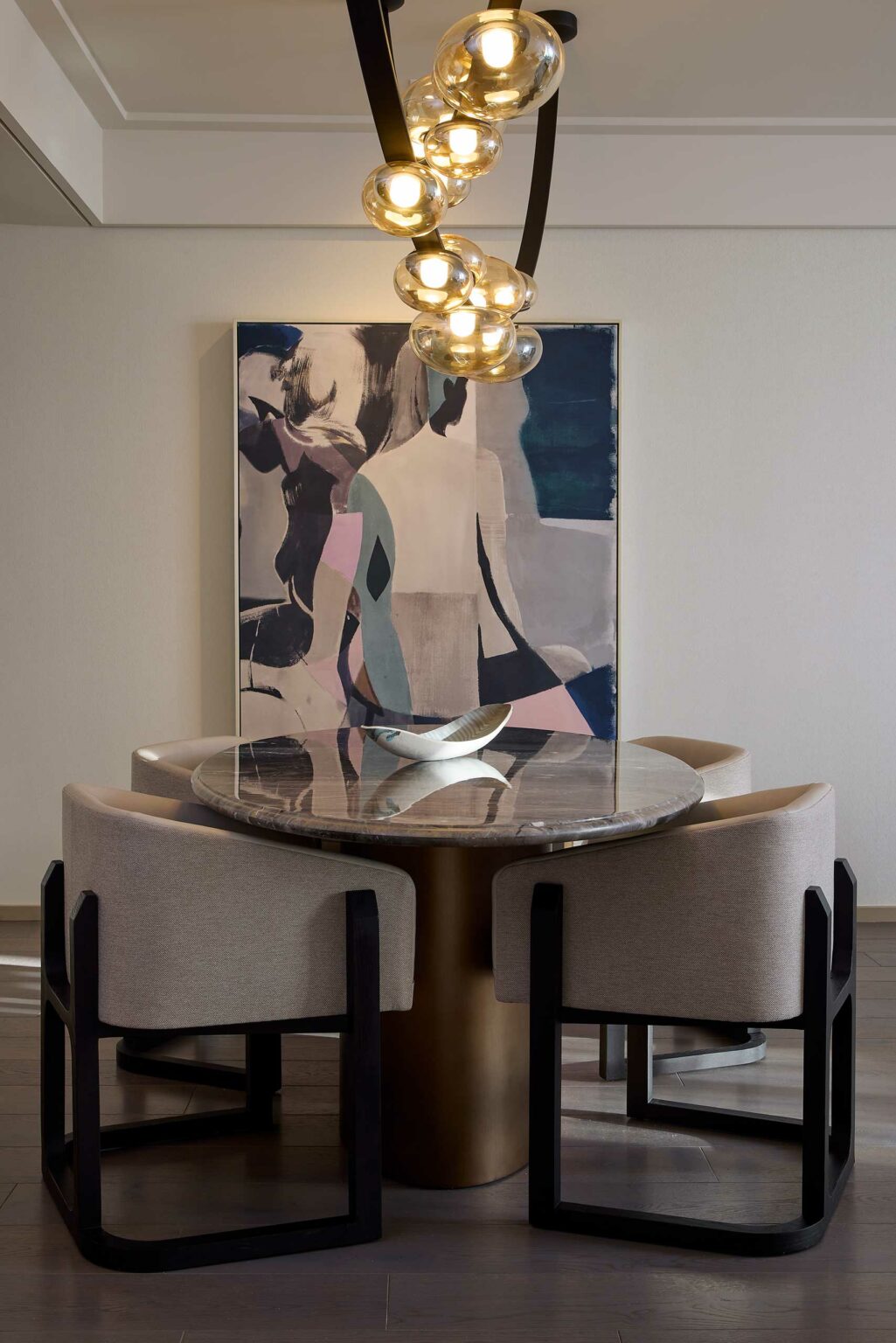 A modern suite dining area features a round marble table with beige chairs, a contemporary painting in the background, and a unique light fixture above.