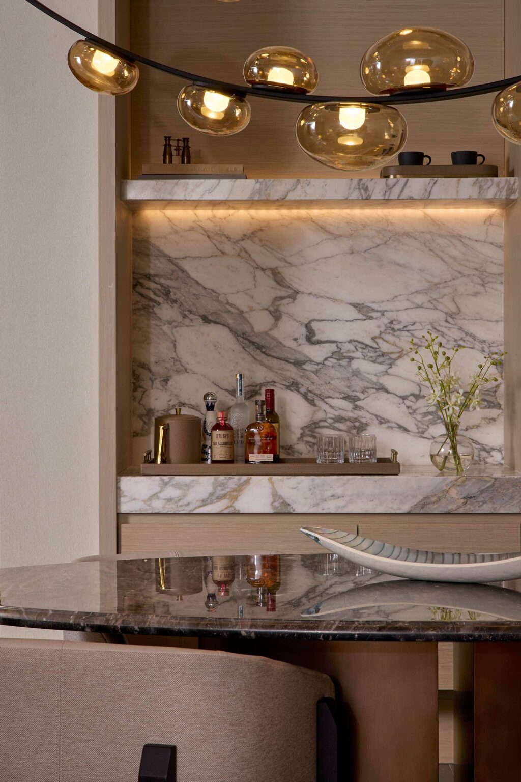 Modern suite kitchen bar area with marble backsplash, amber globe lights, and bottles arranged on a tray. A sleek vase and dish add elegance. Warm, inviting ambiance.