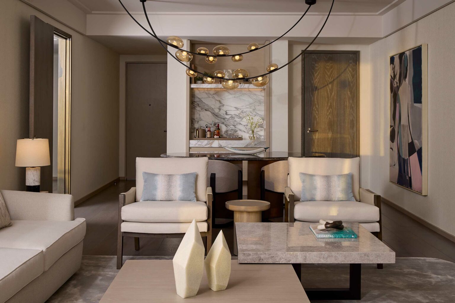 Modern suite living room with neutral tones, featuring beige armchairs and sofa. A marble countertop in the back holds a vase of flowers, while a sleek chandelier hangs above.