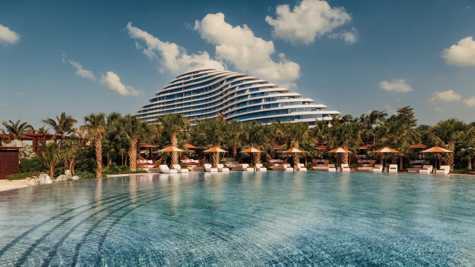 A modern, wave-shaped hotel with curved lines sits behind a palm tree-lined pool. Lounge chairs and orange umbrellas create a relaxing atmosphere.