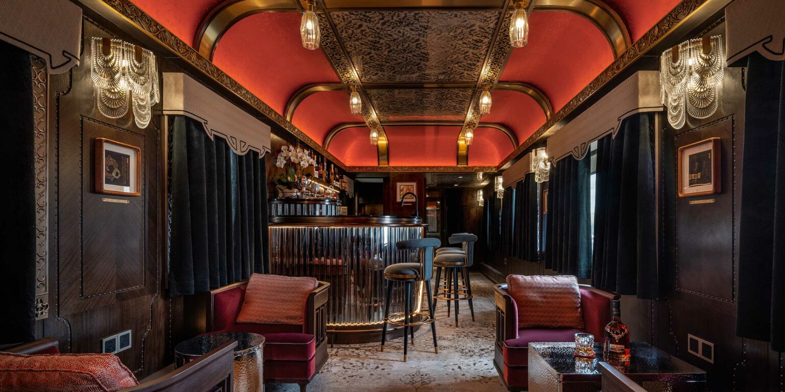 Luxurious vintage train bar with art deco elements. Red ceiling, ornate lighting, plush velvet seats, and polished bar create an elegant ambiance.