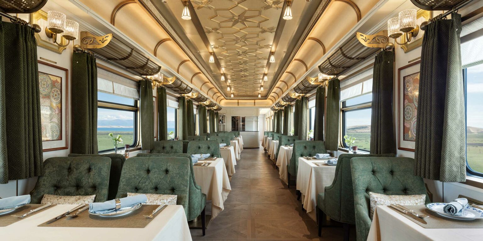 Luxurious train dining car with elegant beige ceiling, geometric patterns, plush green chairs, white tablecloths, and serene atmosphere.