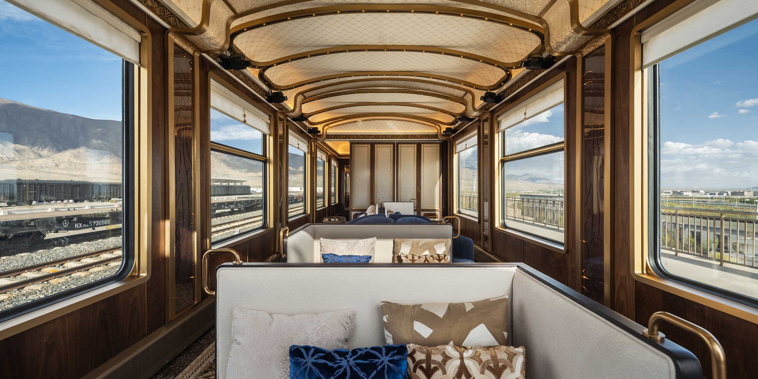 Luxurious train interior with elegant wooden paneling and large windows showing mountain views. Plush seating with decorative pillows creates a cozy atmosphere.