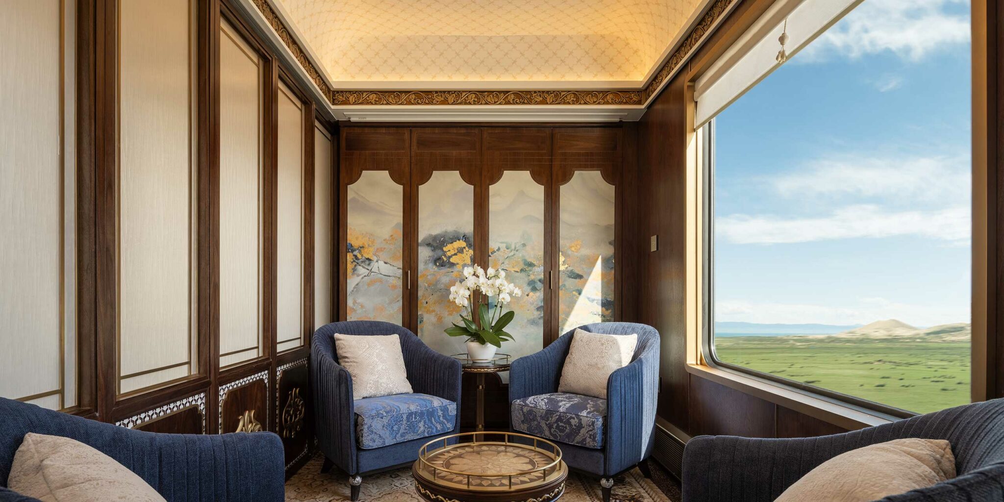 Elegant train cabin with two blue armchairs and a small round table. A potted orchid sits between the chairs. Wall mural depicts serene mountains.