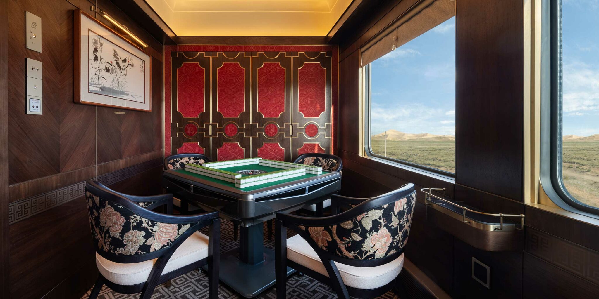 Luxurious train car interior with a mahjong table, floral chairs, and ornate red and gold paneling. Large window shows a scenic landscape, evoking elegance.
