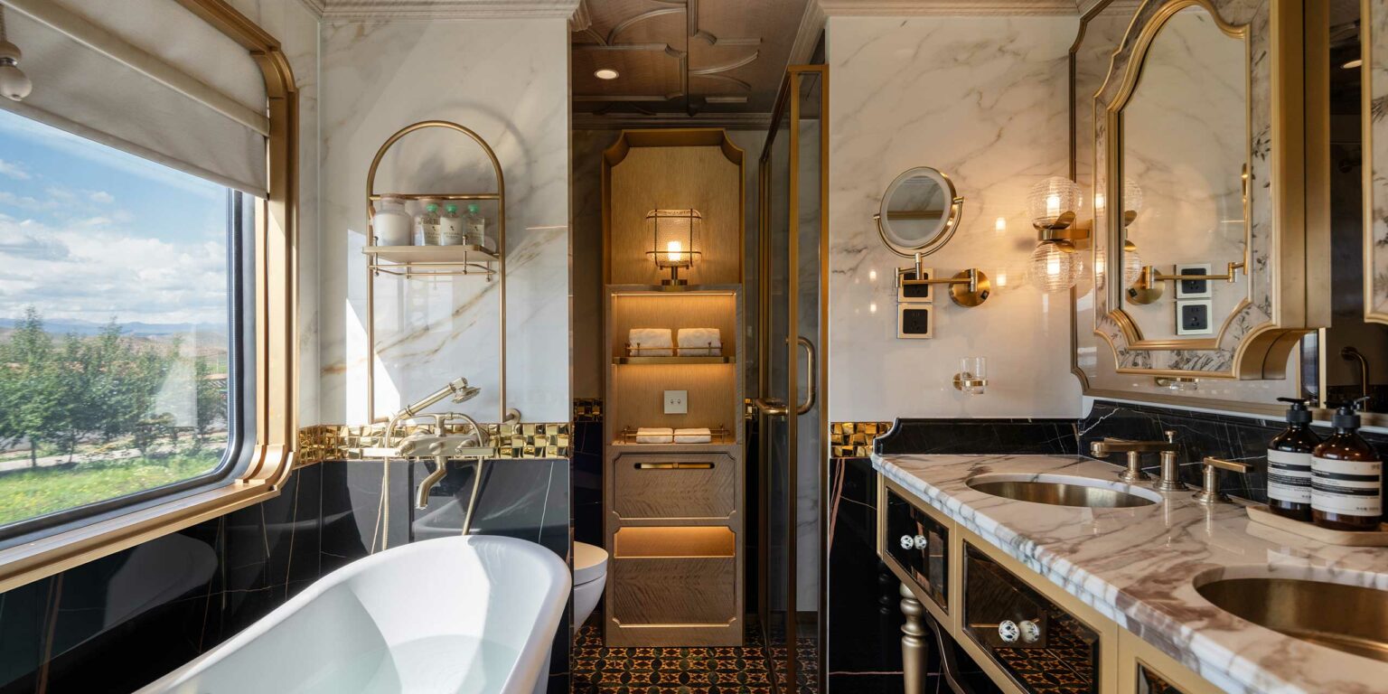 Luxurious train bathroom with elegant marble surfaces, gold fixtures, a freestanding tub, dual sinks, plush towels, and warm lighting. Spacious and opulent.